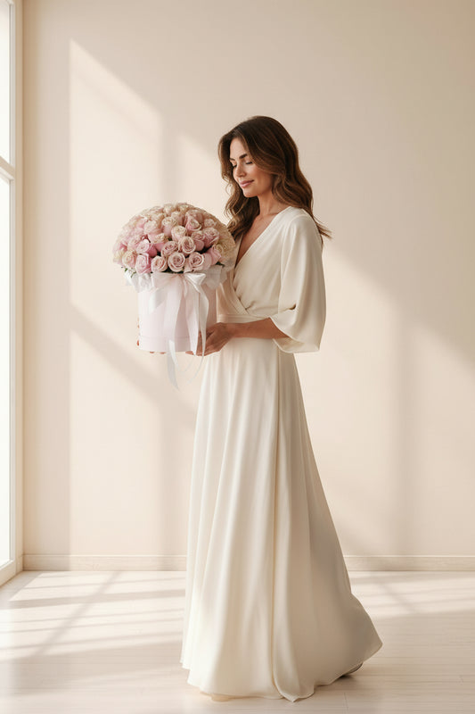 Woman in a white dress holding a box of pink flowers in a bright room.