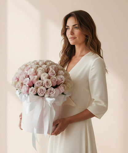 Woman in a white dress holding a box of pink roses in a softly lit room.