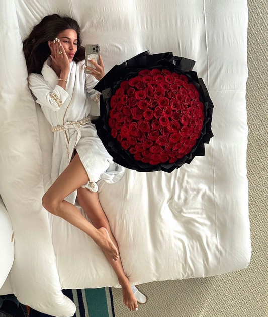 Woman in a white robe lying on a bed next to a large bouquet of red roses.