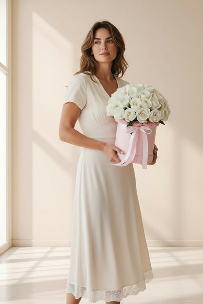Woman in a white dress holding a box of white roses in a softly lit room.