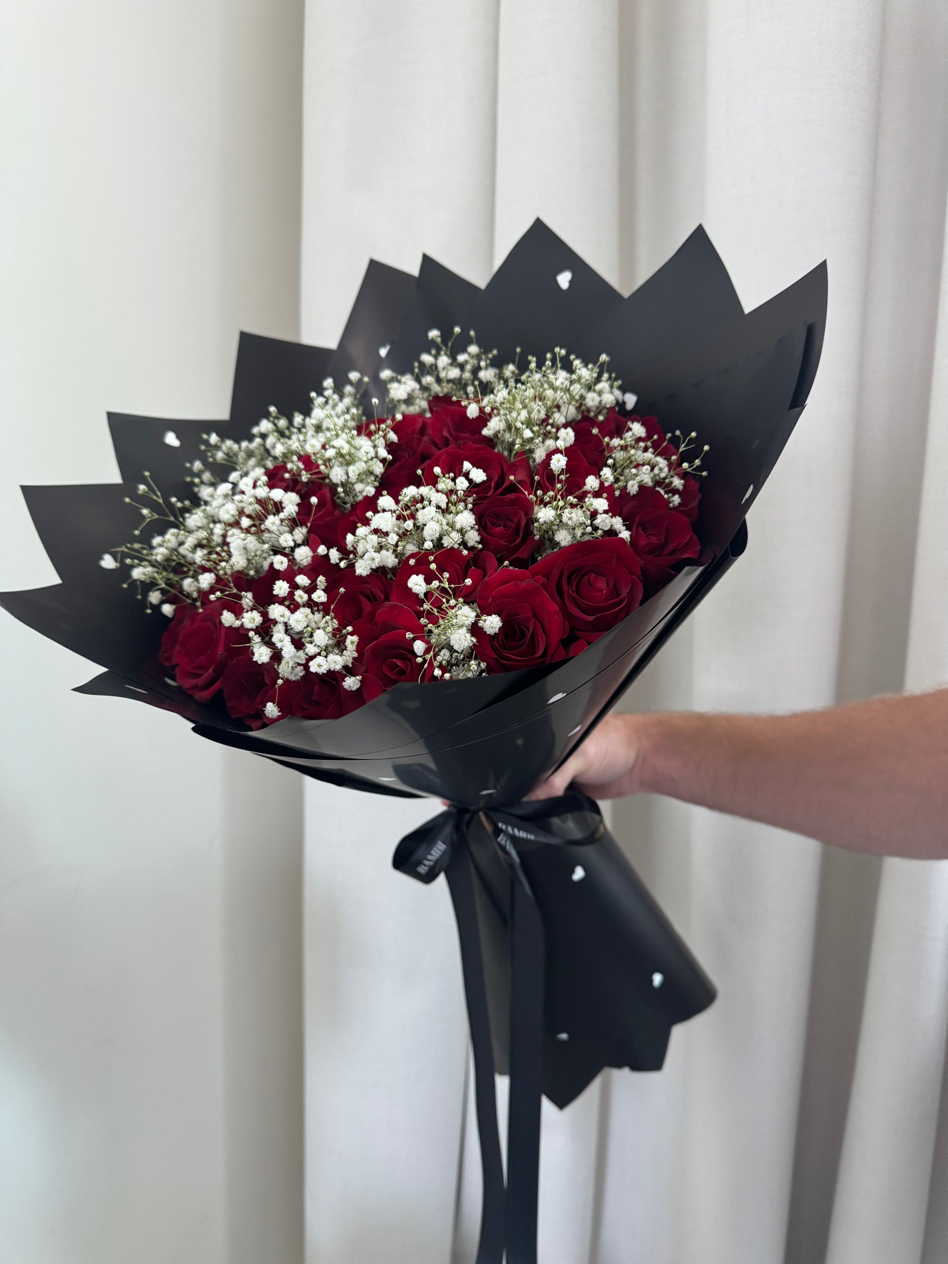 Bouquet of red roses with baby's breath wrapped in black paper, held by a person against a white curtain.
