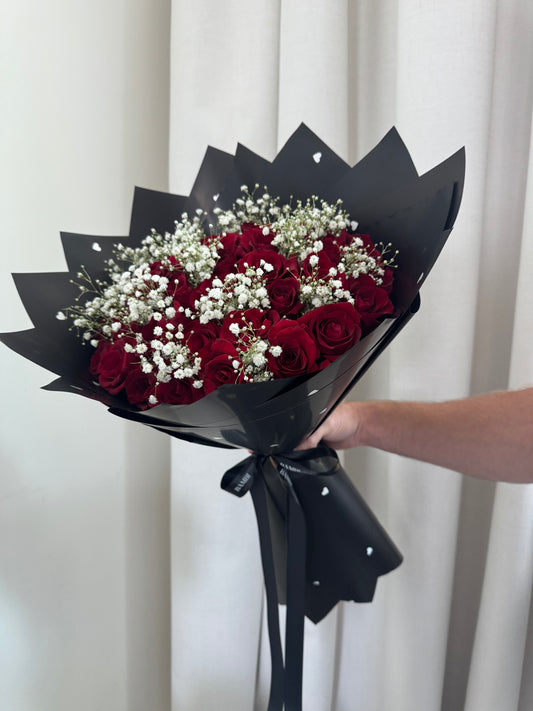 Bouquet of red roses with baby's breath wrapped in black paper, held by a person against a white curtain.