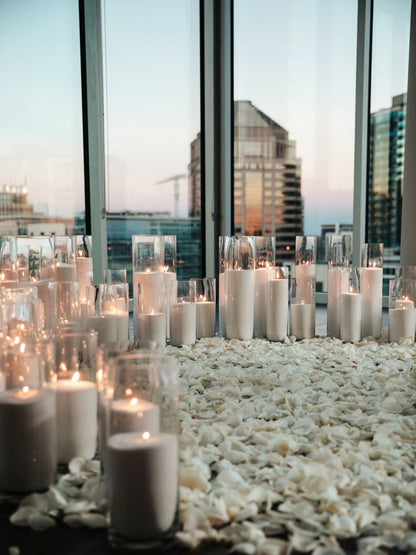 Candlelit setting with candles in glass holders on a pebble surface near large windows with cityscape view.