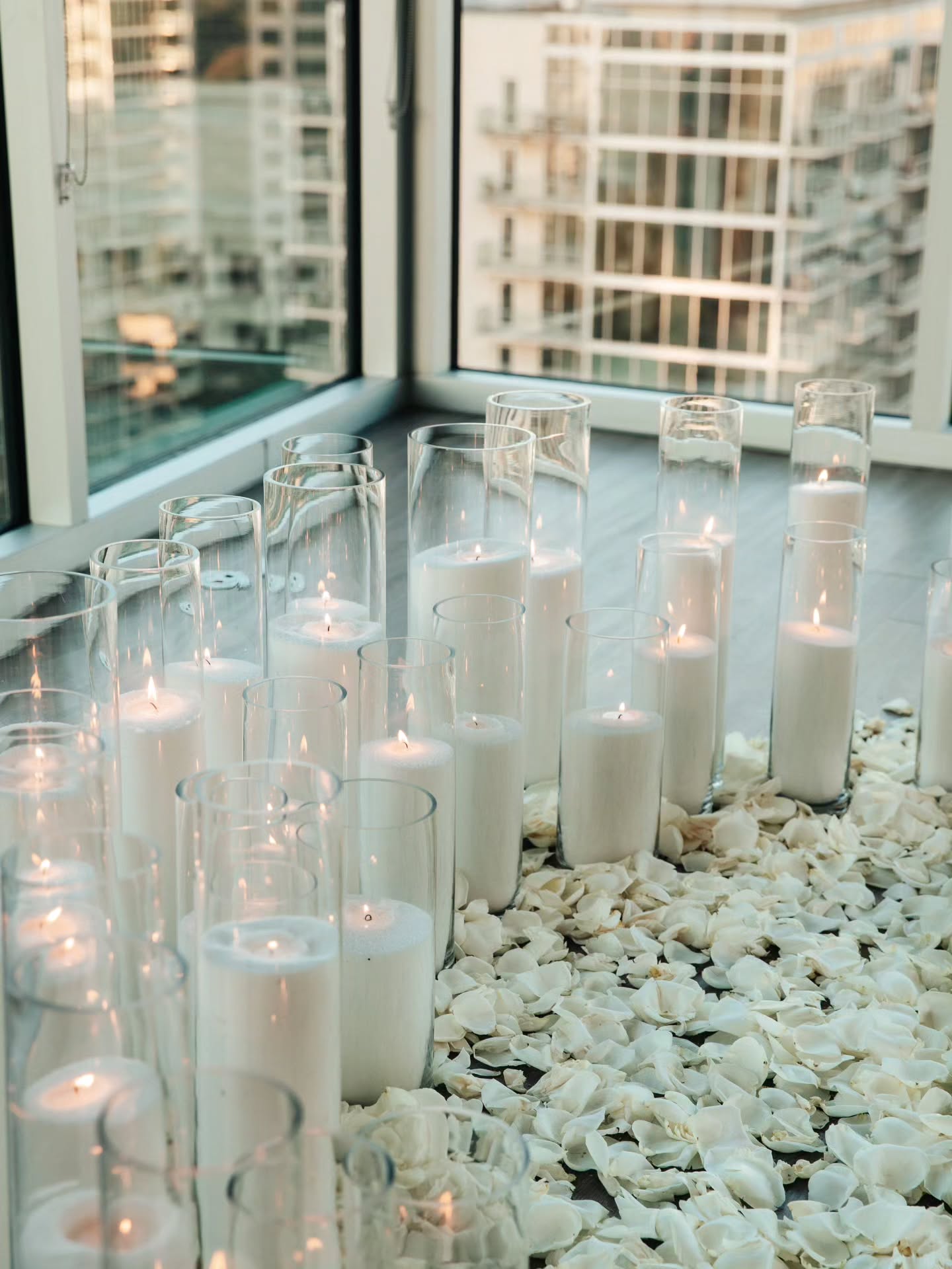 Candlelit setting with white candles in glass holders on a pebble-covered surface near a window.