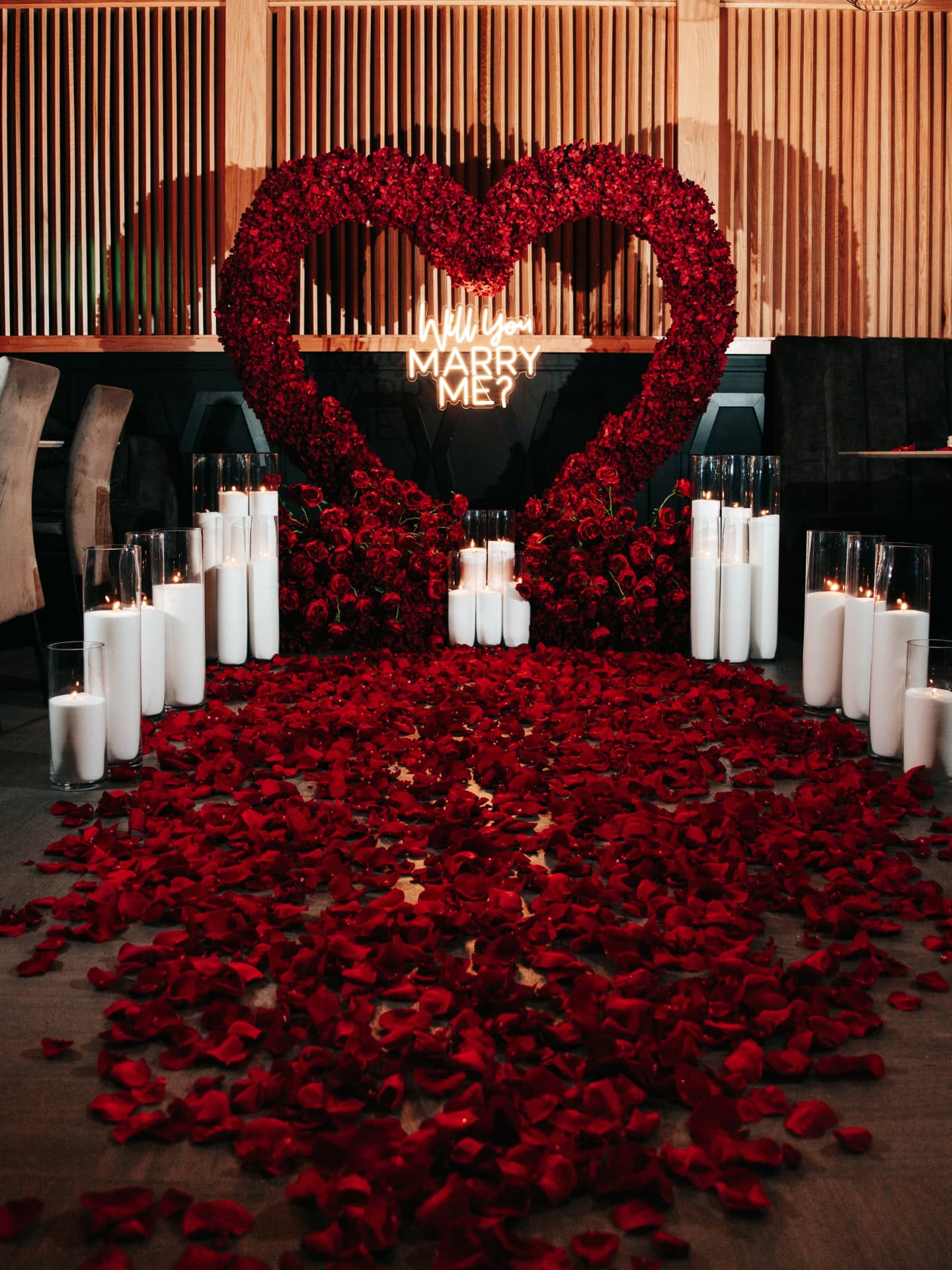Decorative setup with red rose petals, candles, and a heart-shaped arch in a formal setting.