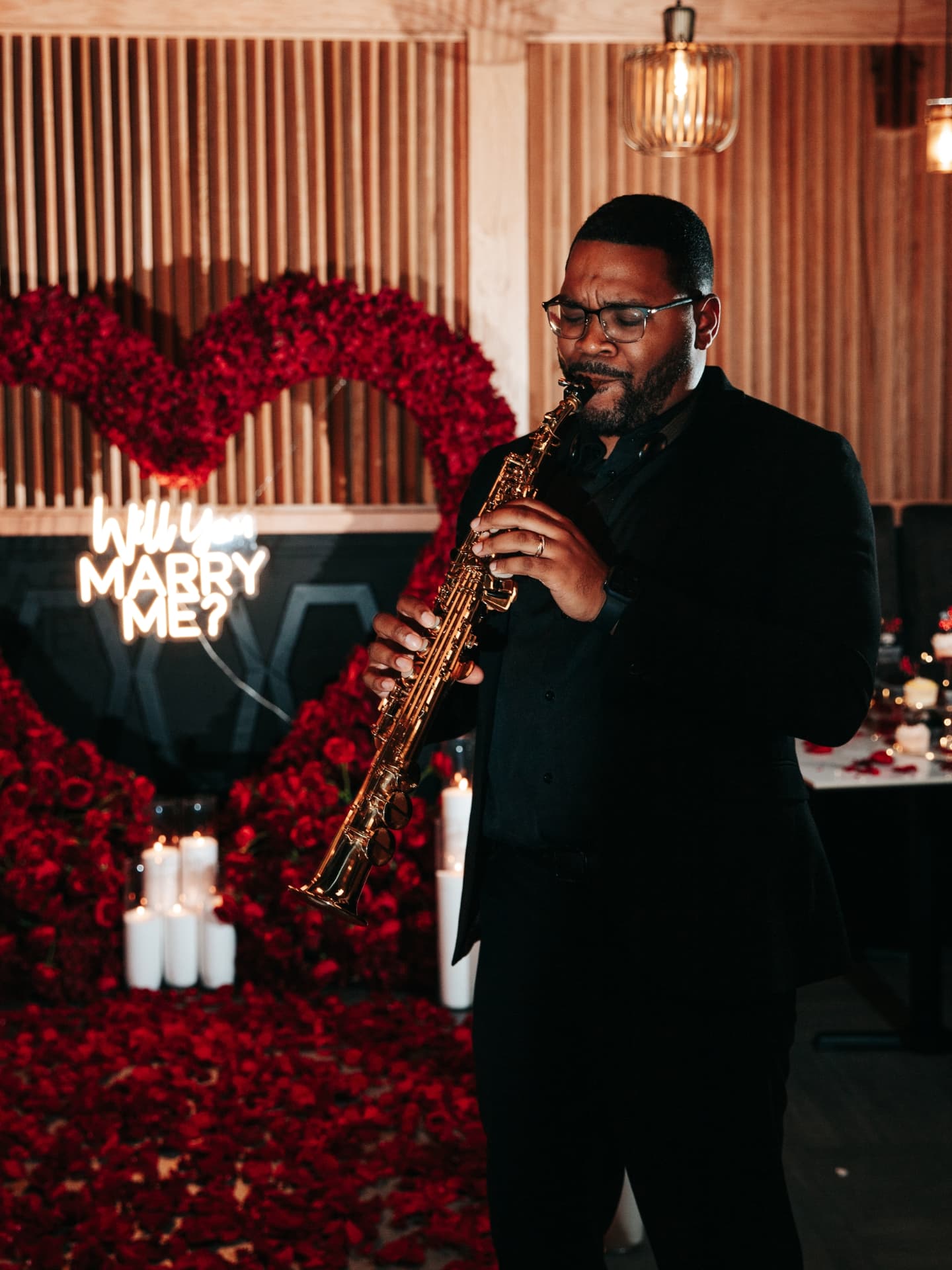 Man playing a saxophone in front of a romantic setup with red roses and 'Will you marry me?' proposal.