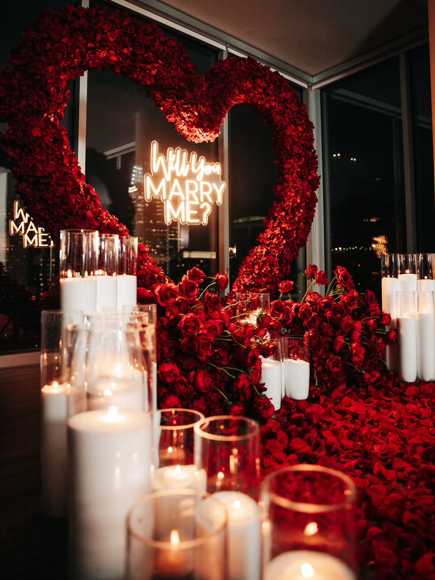 Heart-shaped arrangement of red roses with candles and a 'Will You Marry Me?' neon sign in the background.
