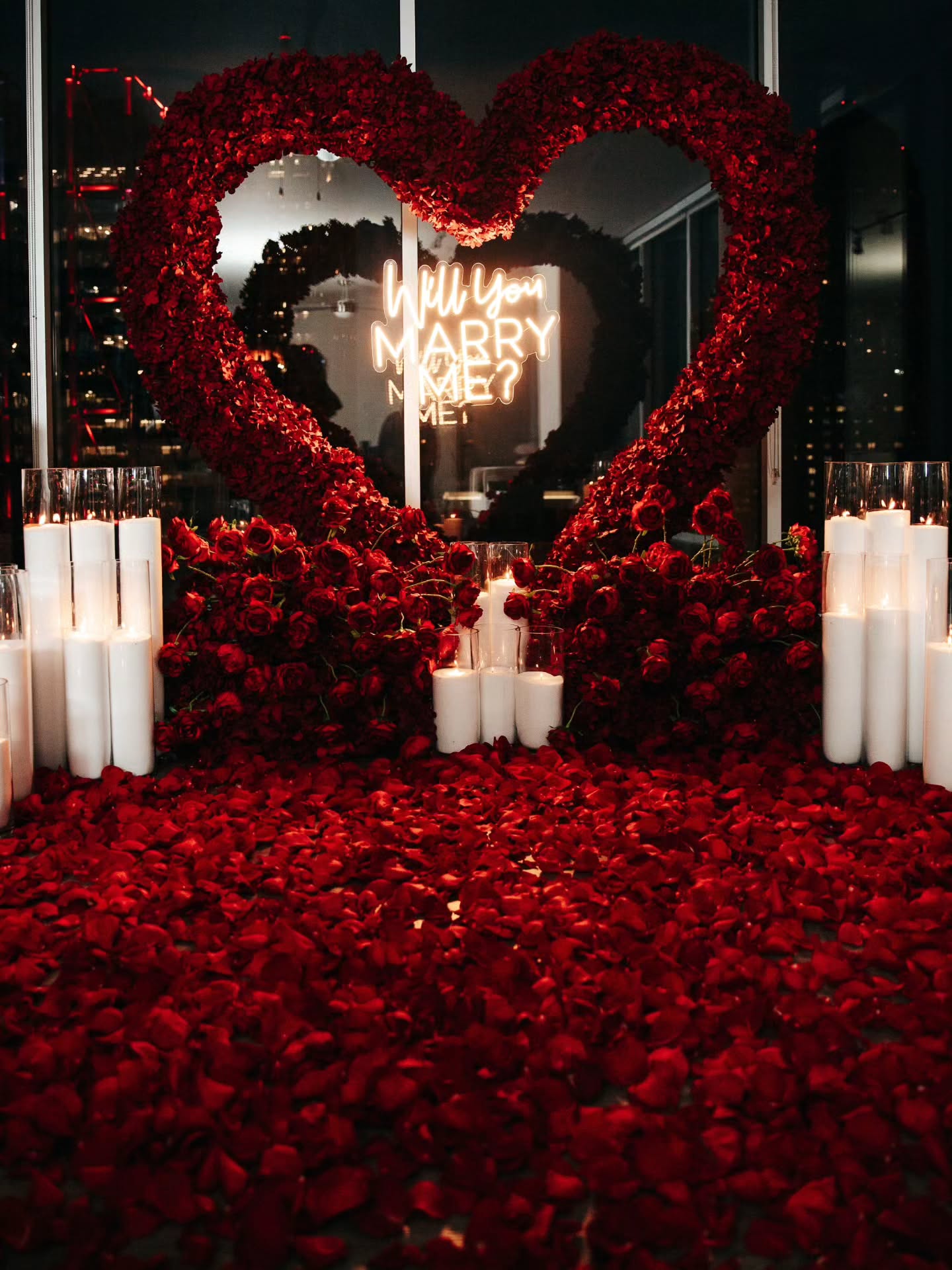 Heart-shaped arrangement of red roses with candles on a dark background, featuring a 'Will You Marry Me?' proposal.