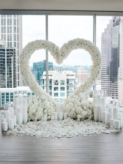 Heart-shaped arrangement of white flowers with candles in a room with large windows showing cityscape.