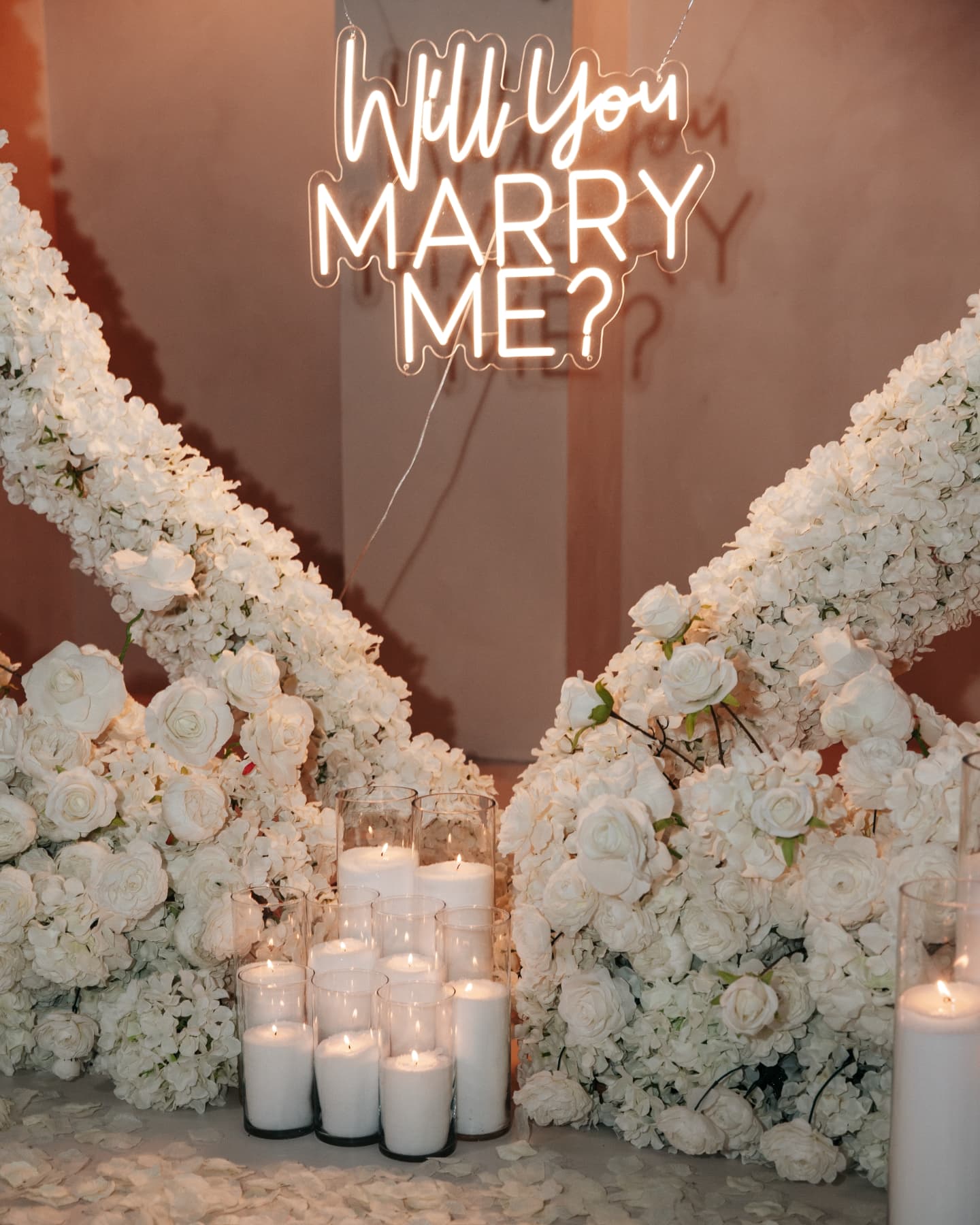 Neon sign asking 'Will You Marry Me?' surrounded by white flowers and candles.