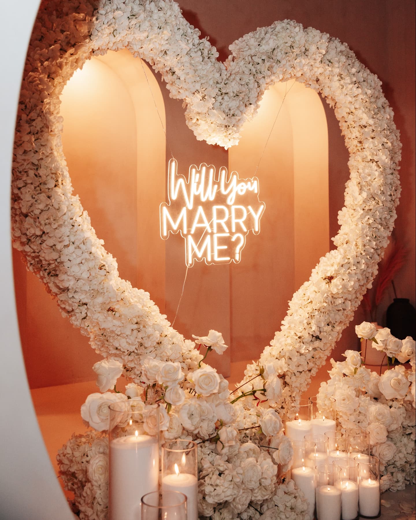 Heart-shaped floral arrangement with 'Will You Marry Me?' neon sign and candles on a warm-toned background
