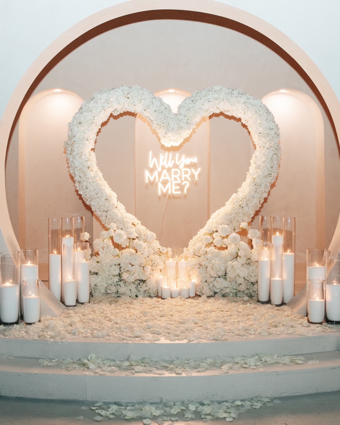 Heart-shaped floral arrangement with 'Will You Marry Me?' neon sign and candles on a romantic setting.