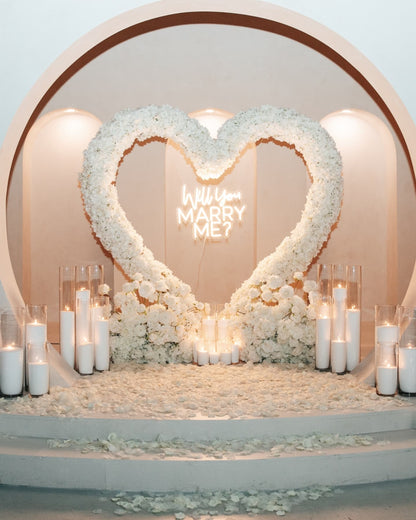 Heart-shaped floral arrangement with 'Will You Marry Me?' neon sign and candles on a romantic setting.