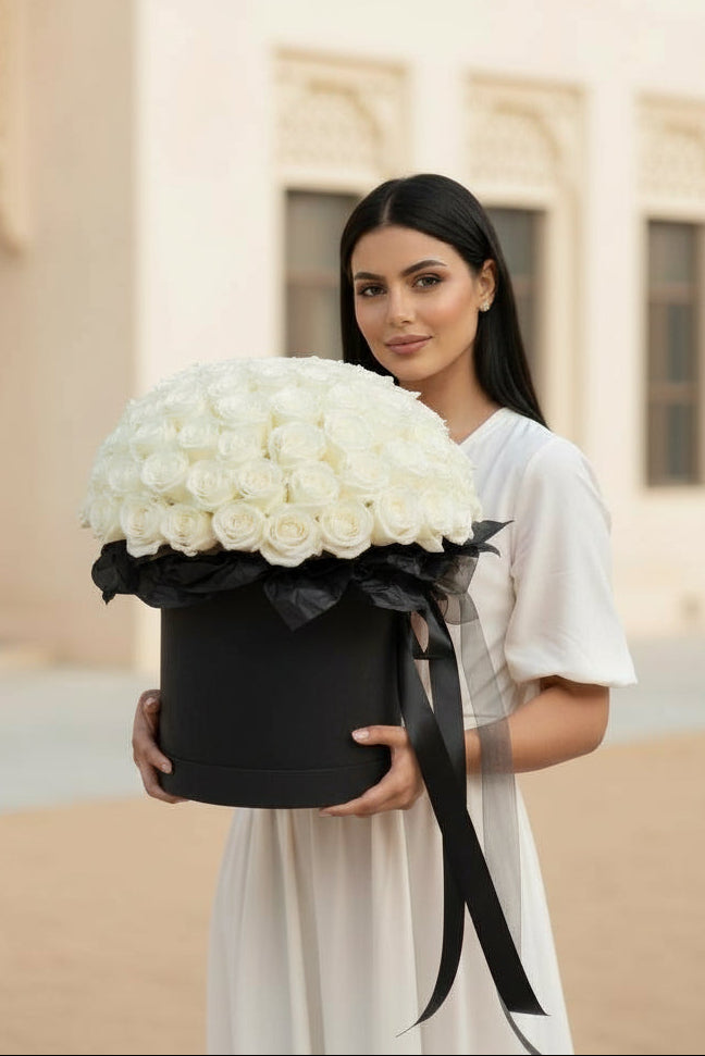 Woman holding a large black box of white flowers in front of a beige building.