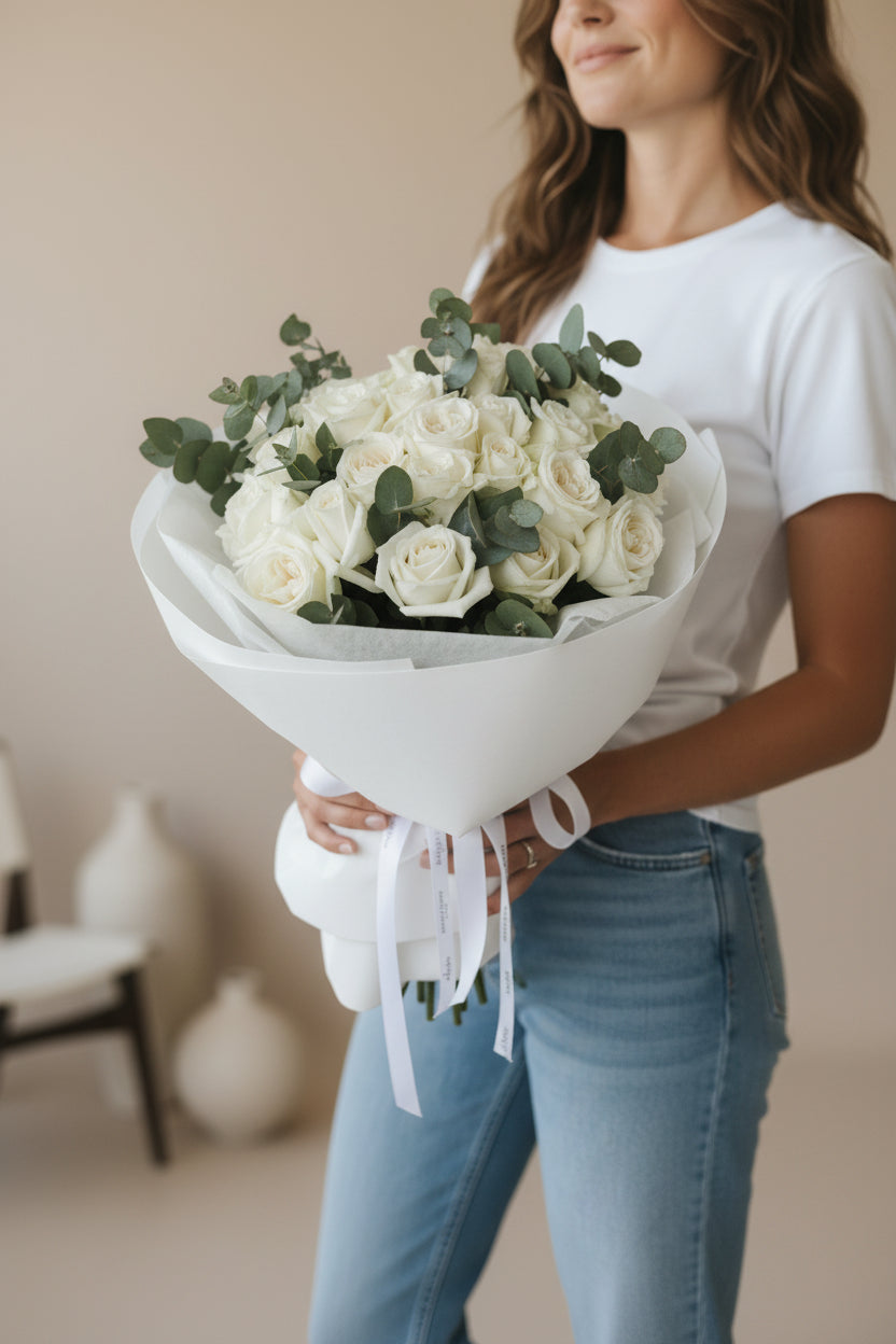 Person holding a bouquet of white roses and greenery in a neutral setting