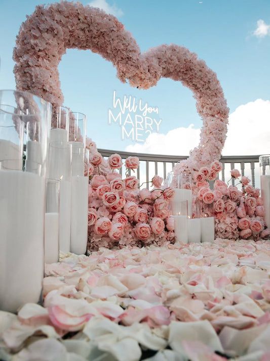 Romantic setup with pink floral arch and 'Will You Marry Me' proposal card.