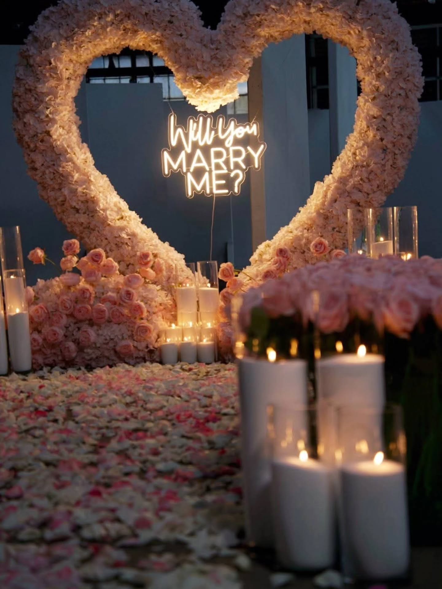 Floral heart arrangement with 'Will You Marry Me?' light sign and candles on a romantic setting.
