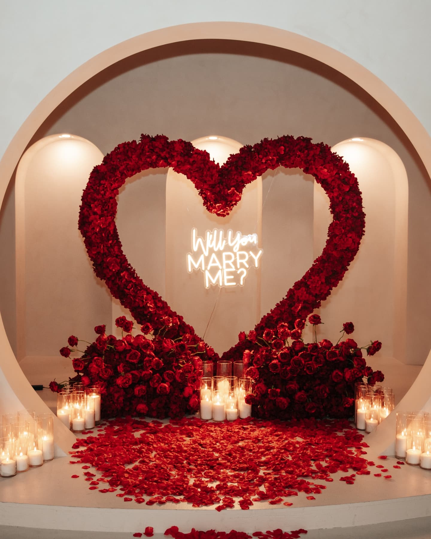 Heart-shaped arrangement of red roses with 'Will You Marry Me?' neon sign on a romantic setup.