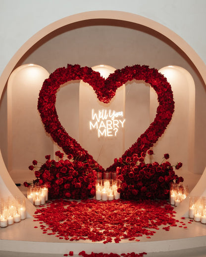 Heart-shaped arrangement of red roses with 'Will You Marry Me?' neon sign on a romantic setup.