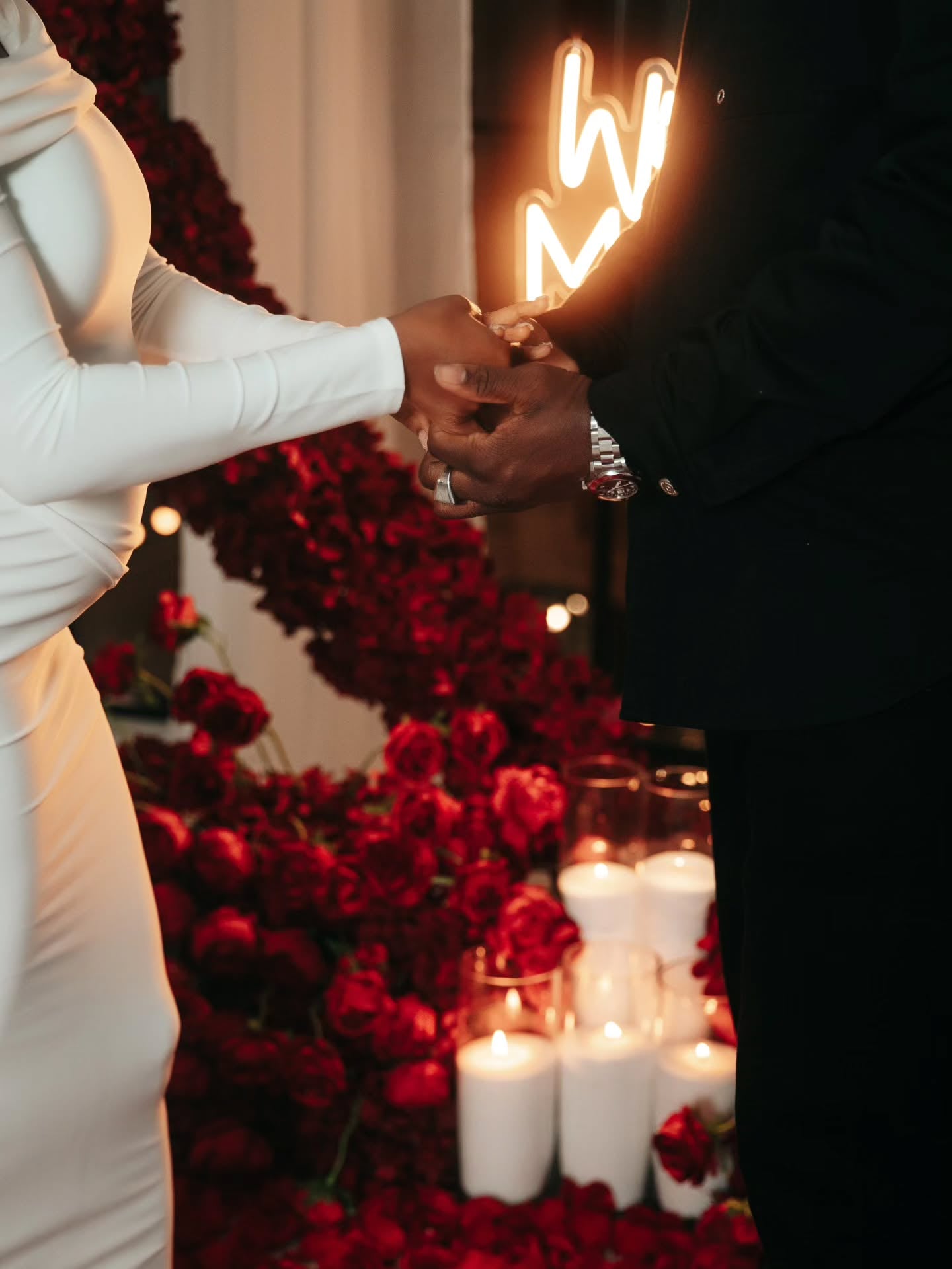 Two people holding hands with a heart-shaped light bulb above, surrounded by red flowers and candles.