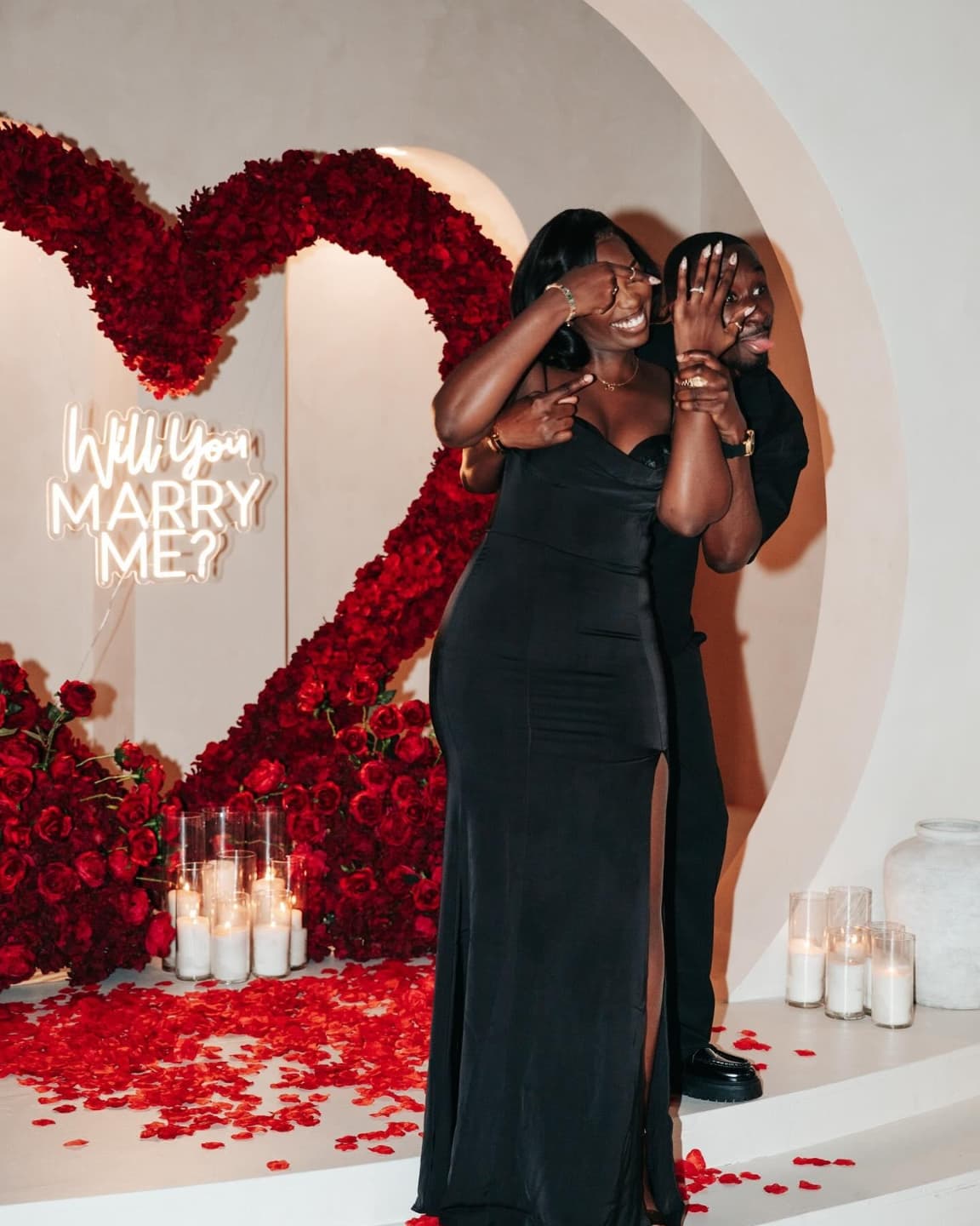 Woman in a black dress standing in front of a heart-shaped floral arrangement with 'Will You Marry Me?' proposal.