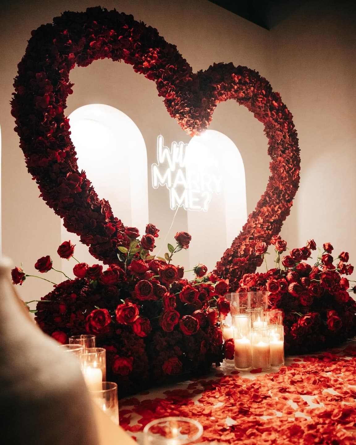 Heart-shaped floral arrangement with candles on a red surface, featuring the text 'Will you marry me' in the background.