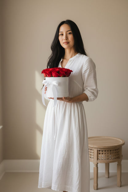 Woman in a white dress holding a box of red roses against a beige wall.