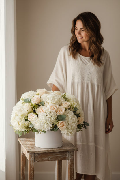 Woman in a white dress standing next to a large floral arrangement on a wooden table.