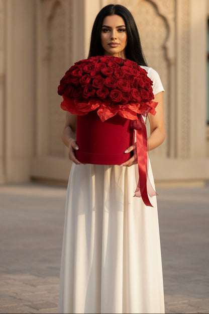 Woman holding a large bouquet of red roses in a decorative box against an architectural background.