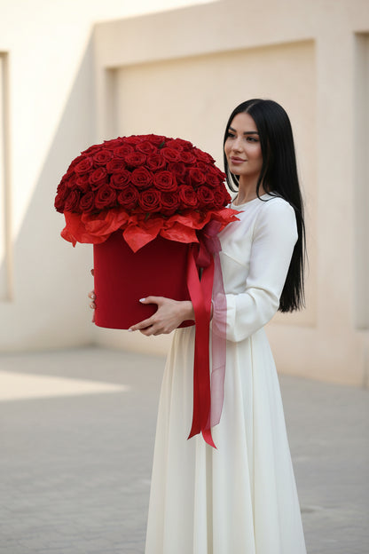 Woman holding a large bouquet of red roses in a decorative box with a red ribbon against a light background.