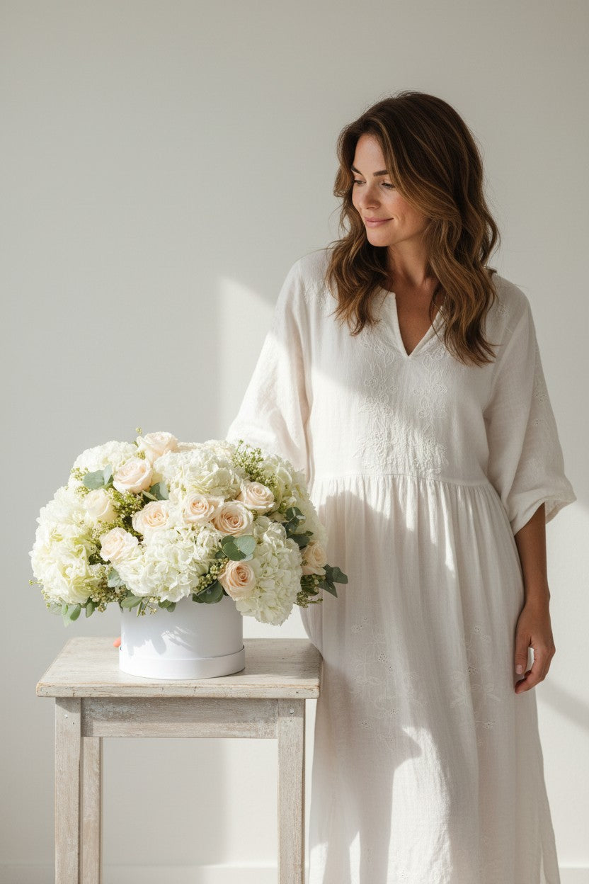 Woman in a white dress standing next to a table with a floral arrangement against a plain background