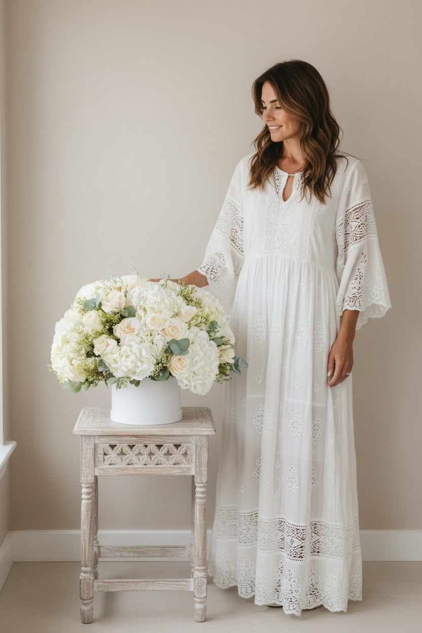 Woman in a white lace dress standing next to a table with a floral arrangement in a neutral setting