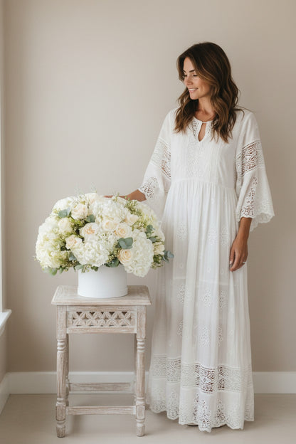 Woman in a white lace dress standing next to a table with a floral arrangement in a neutral setting